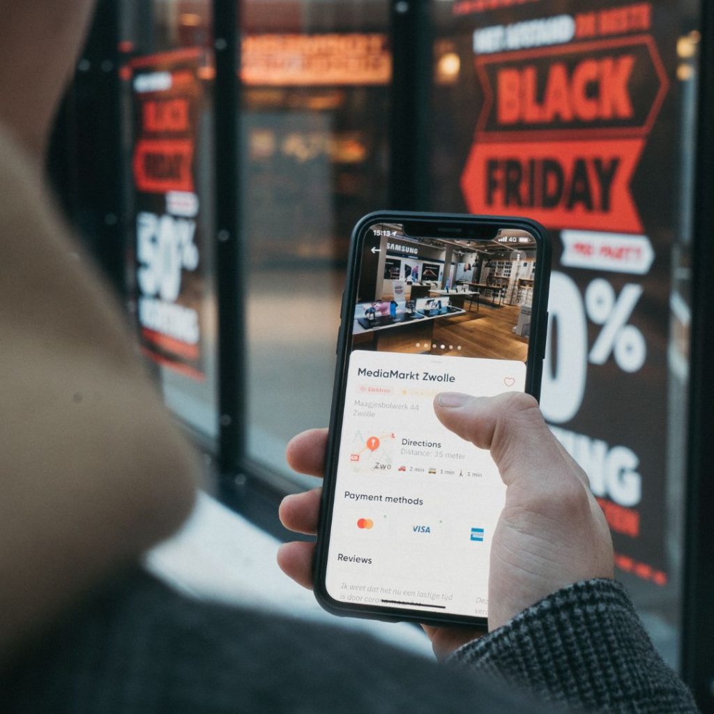 Smartphone displaying a digital commerce app, placed in front of a physical store with Black Friday signage, illustrating the importance of digital commerce in modern retail
